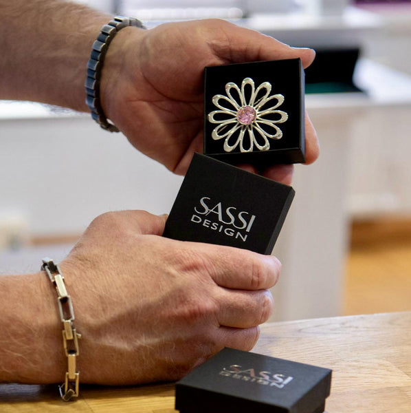 Lähikuva Sassi Designin käsintehdystä hopeisesta rintakorusta “Kesän Kukka”. Kauniisti muotoiltu kukka, jossa vaaleanpunainen kivi keskellä, tuo koruun elämän ja värin. Tämä koru on täydellinen lahja itselle tai tärkeälle ihmiselle – tyylikäs ja ainutlaatuinen.
